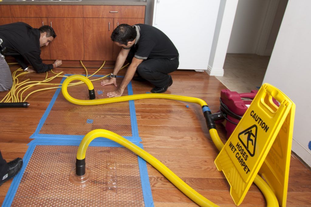 Two men are working on a wooden floor next to a yellow caution sign.