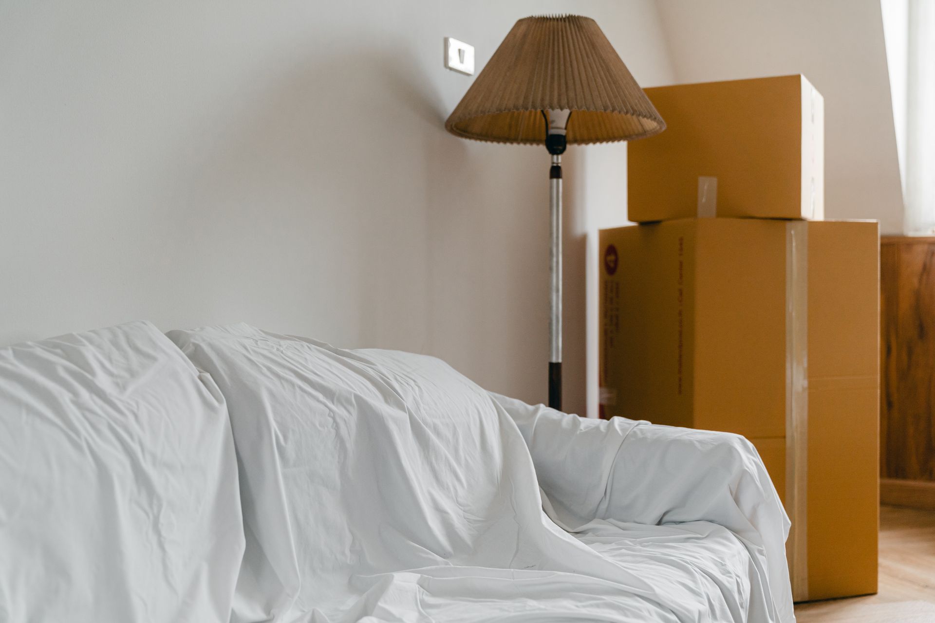 A couch is covered in a white blanket in a room with cardboard boxes and a lamp.