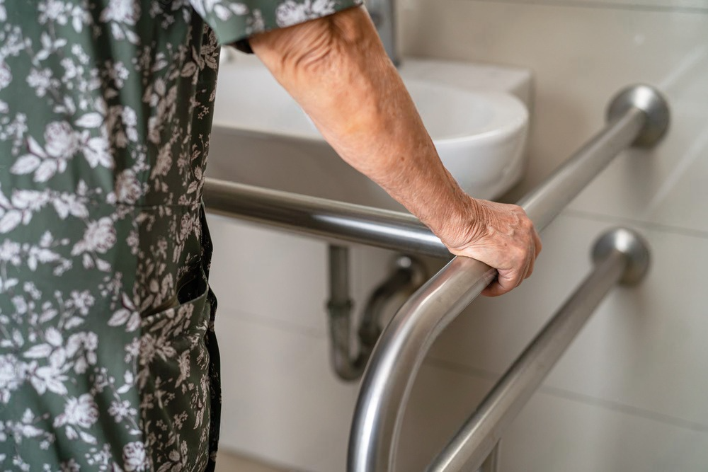An elderly woman is holding onto a metal railing in a bathroom.