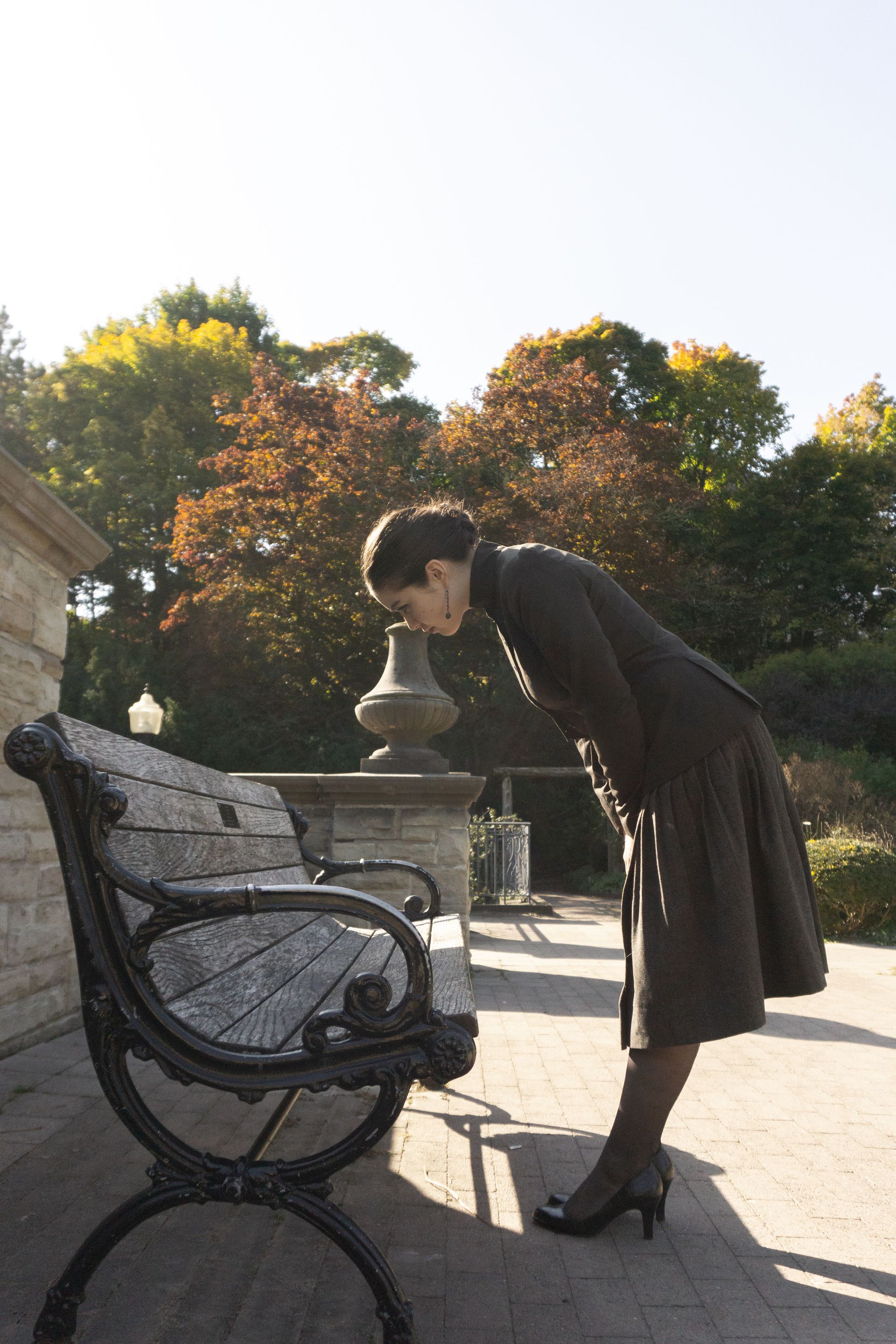 A woman in a suit leaning over to read the inscription on a park bench in autumn.