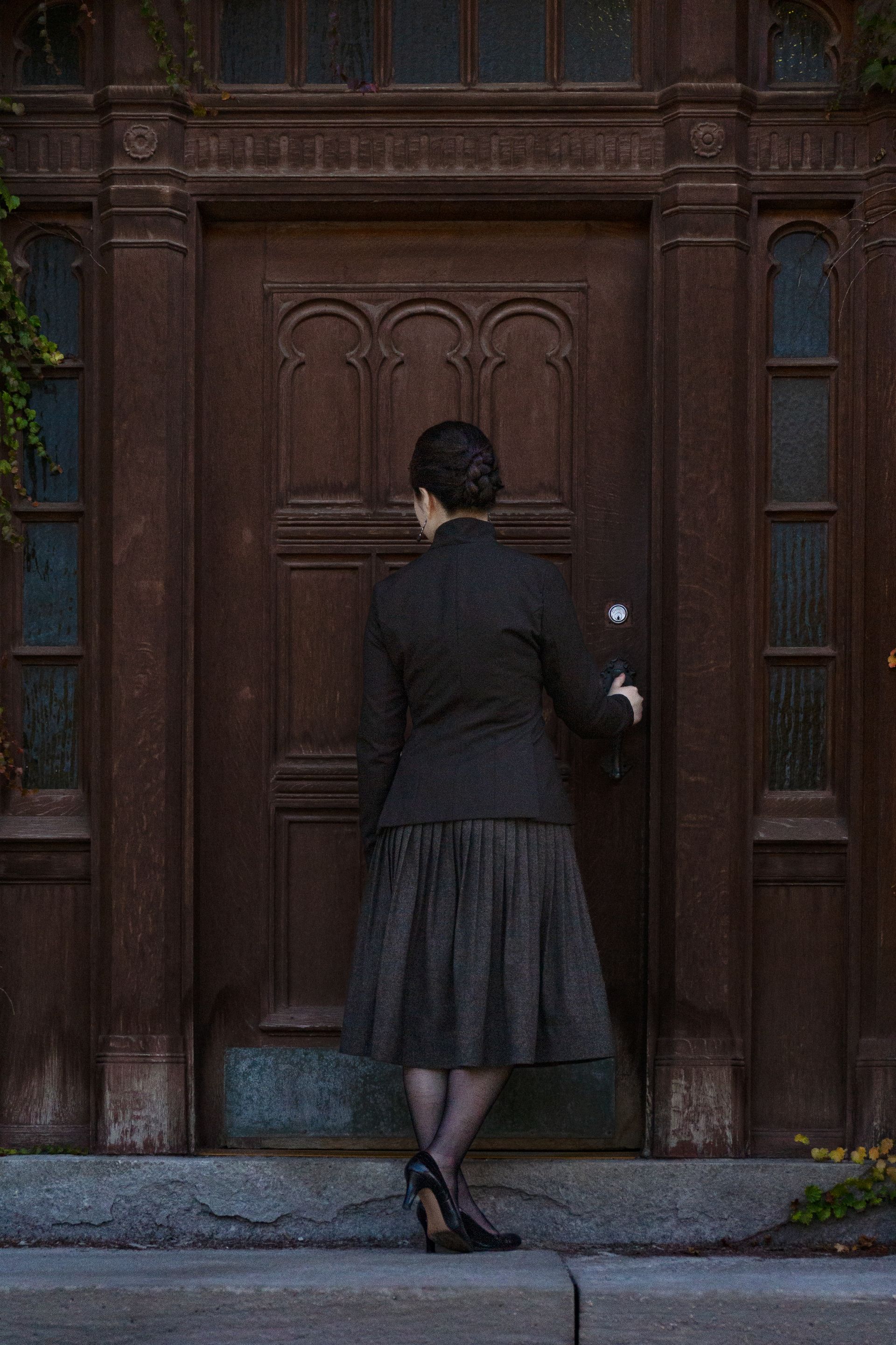 A woman in a suit with her back turned standing in front of an old wooden door.