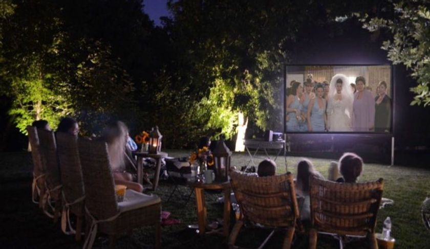 People watching a movie outdoors at night on a large screen. Chairs, snacks, and lanterns are visible.