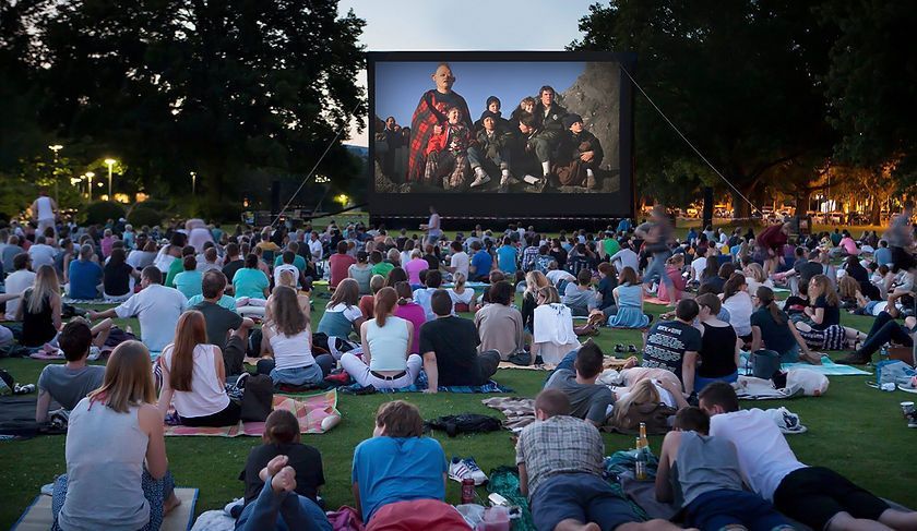 Outdoor movie night; crowd seated on grass watching a film on a large screen at dusk.