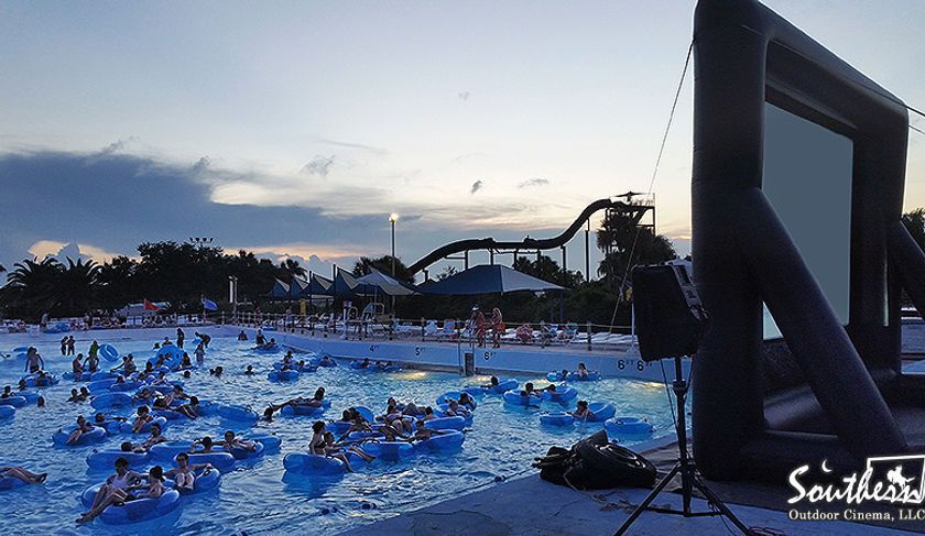 People in inner tubes watch a movie on a screen at a water park at dusk.
