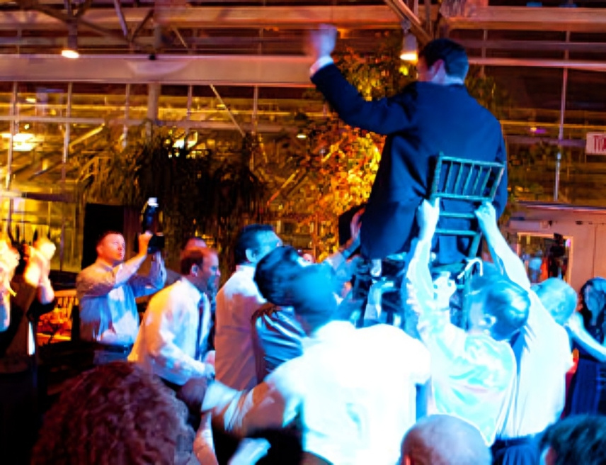 Man in suit on a chair held aloft by a crowd at a celebration with colorful lighting.