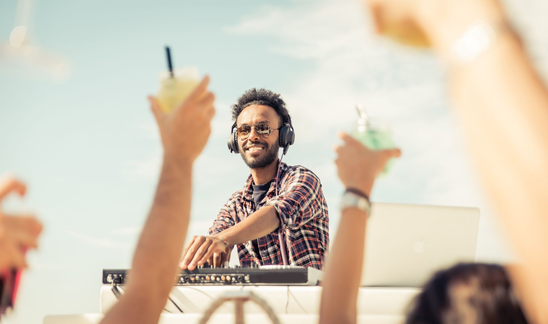 DJ with headphones smiles as people raise drinks in the sky.