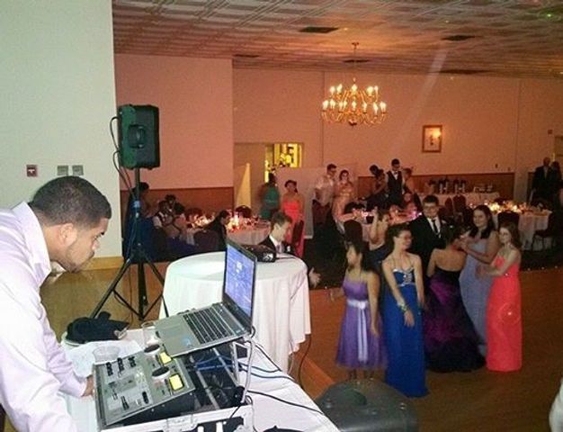DJ at a formal event; people dancing. DJ in pink shirt, laptop, mixing board. Ballroom with chandelier.