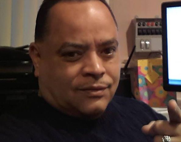 Man pointing, looking at the viewer. He's in a room, a monitor is visible.