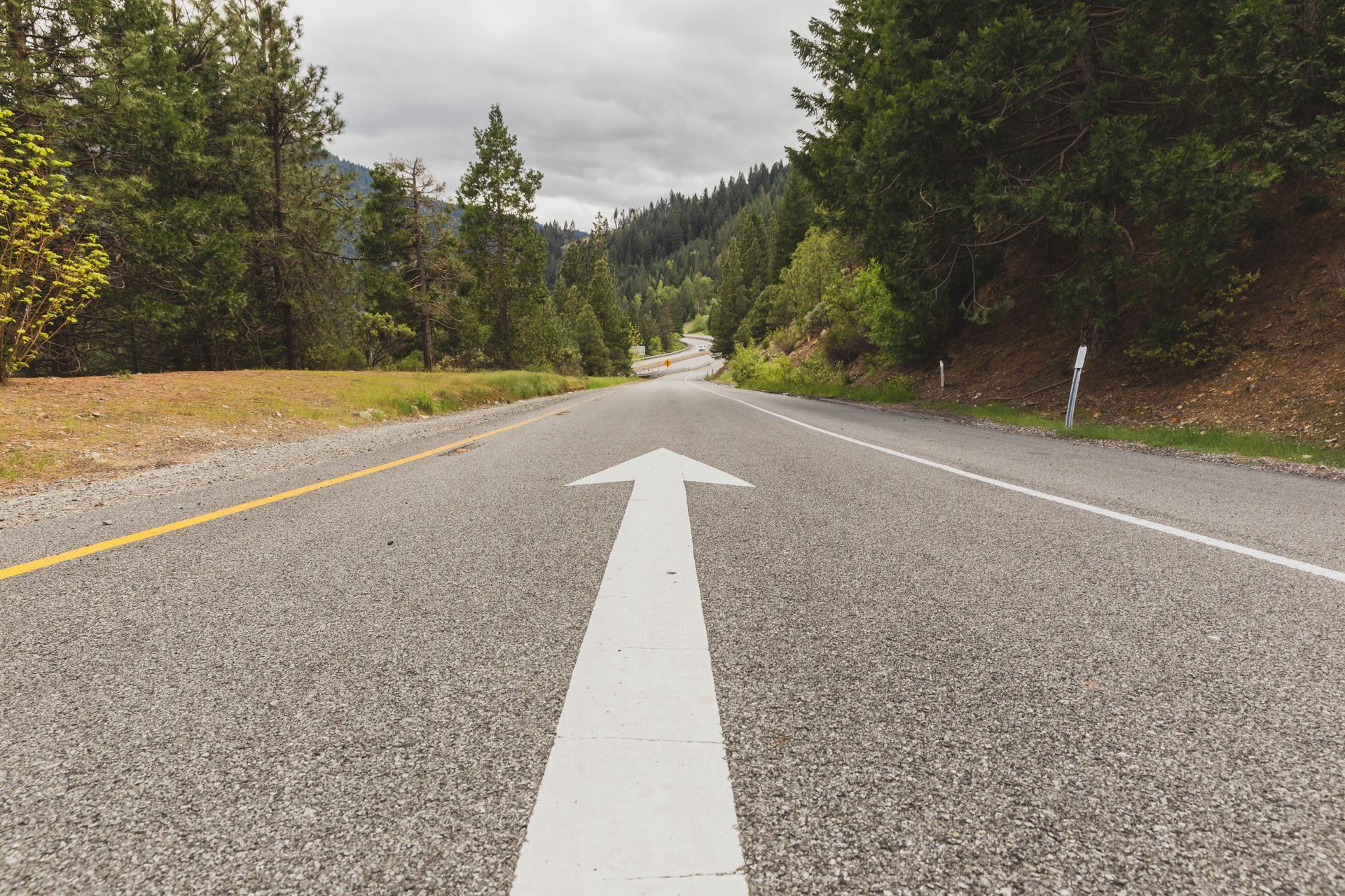 Paved road with a large white arrow, flanked by trees and leading into a mountain range under a cloudy sky.