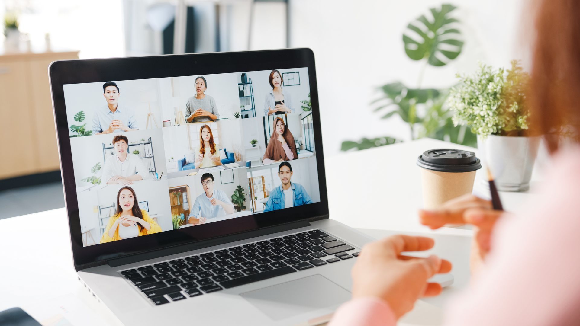 Woman in a video conference with a laptop, looking at the screen. Other people appear in the video, inside a bright room.