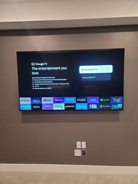 Wall-mounted TV displaying Google TV setup screen, with app icons along the bottom. Brown wall, white trim.
