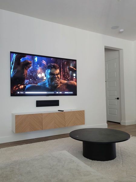 A wall-mounted TV displaying a scene featuring the Hulk, above a floating media console; black coffee table and white rug.