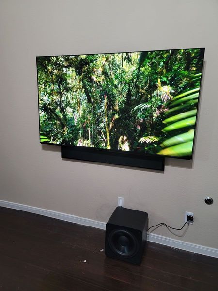 Large TV mounted on a tan wall with a soundbar below. A subwoofer sits on the floor beneath.