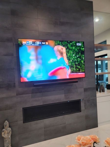 TV on a dark tiled wall above a fireplace, displaying a sports game. Soundbar below TV.