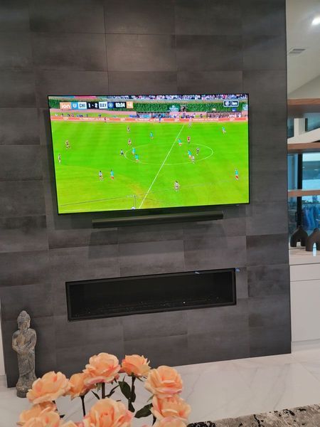 TV displaying a soccer match, mounted on a gray tiled wall above a fireplace, flowers in the foreground.