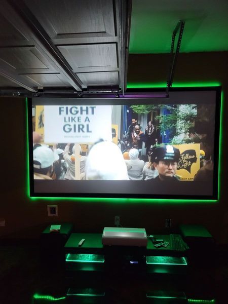 Movie screen in a home theater with green neon lights. A projector displays a scene with a protest sign that reads