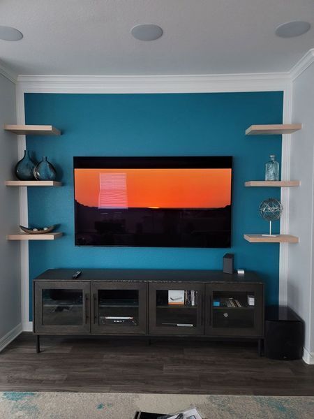 Teal accent wall with floating shelves, TV, and console. Orange and black image displayed. Wooden floor and white trim.