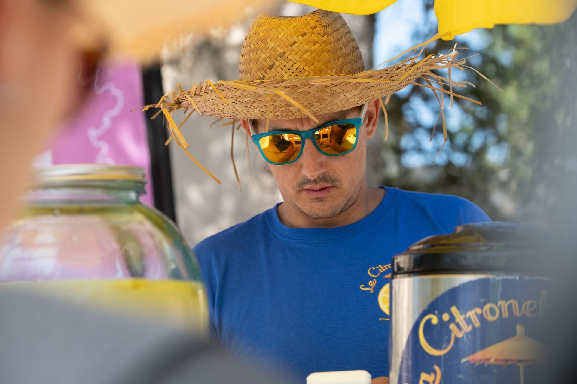 Un homme portant un chapeau de paille et des lunettes de soleil vend de la limonade.