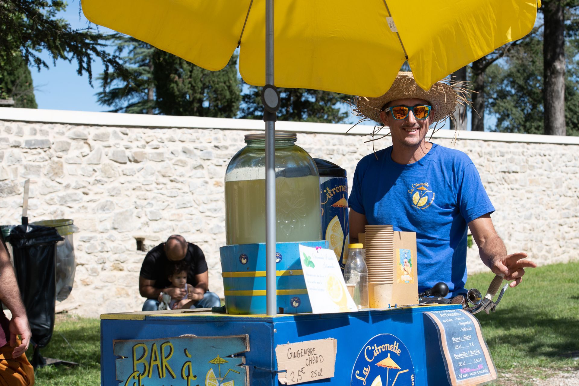 Un homme se tient devant un stand de limonade avec un parapluie jaune.