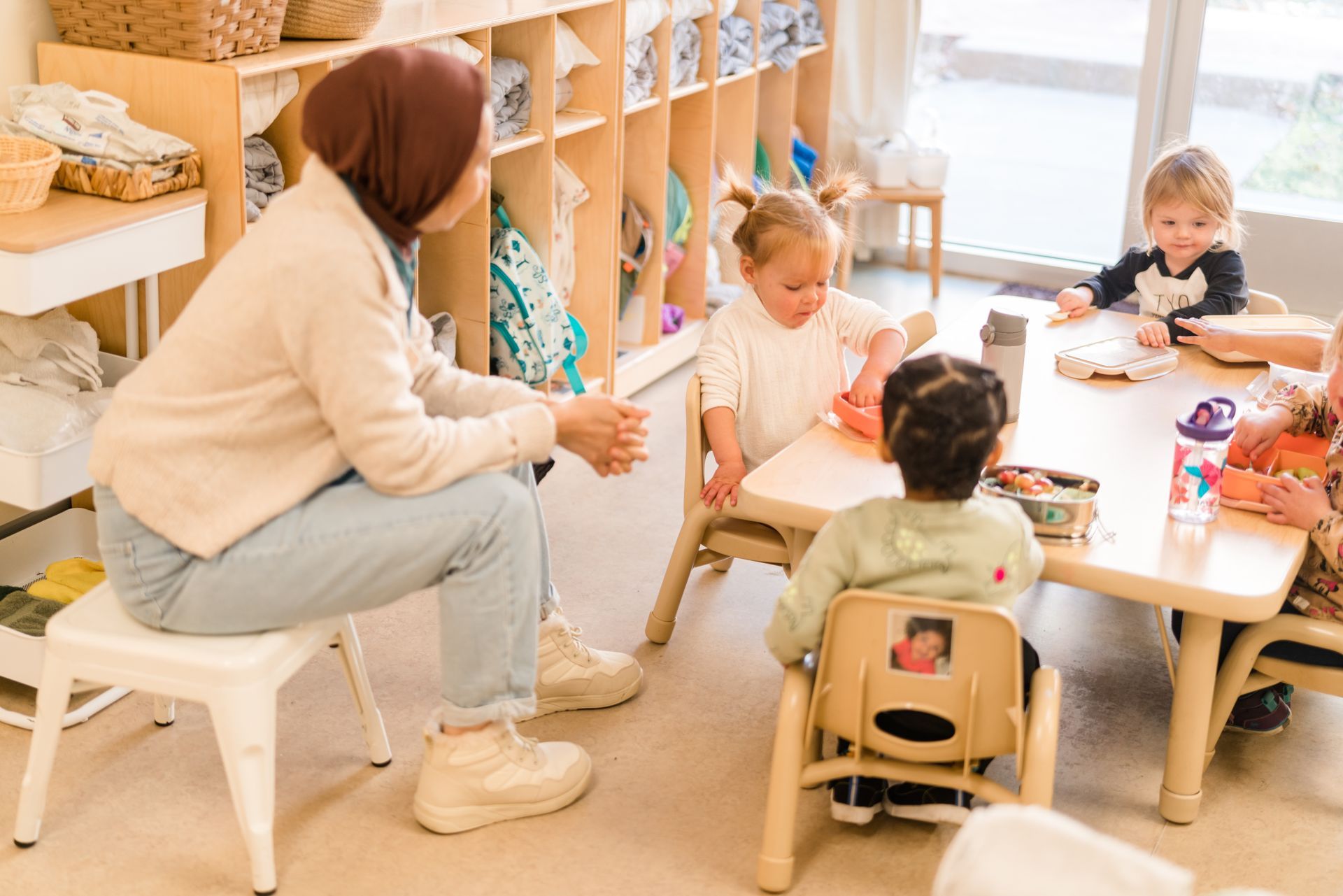 A montessori guide is sitting on a stool talking to a group of children sitting at tables in a classroom.