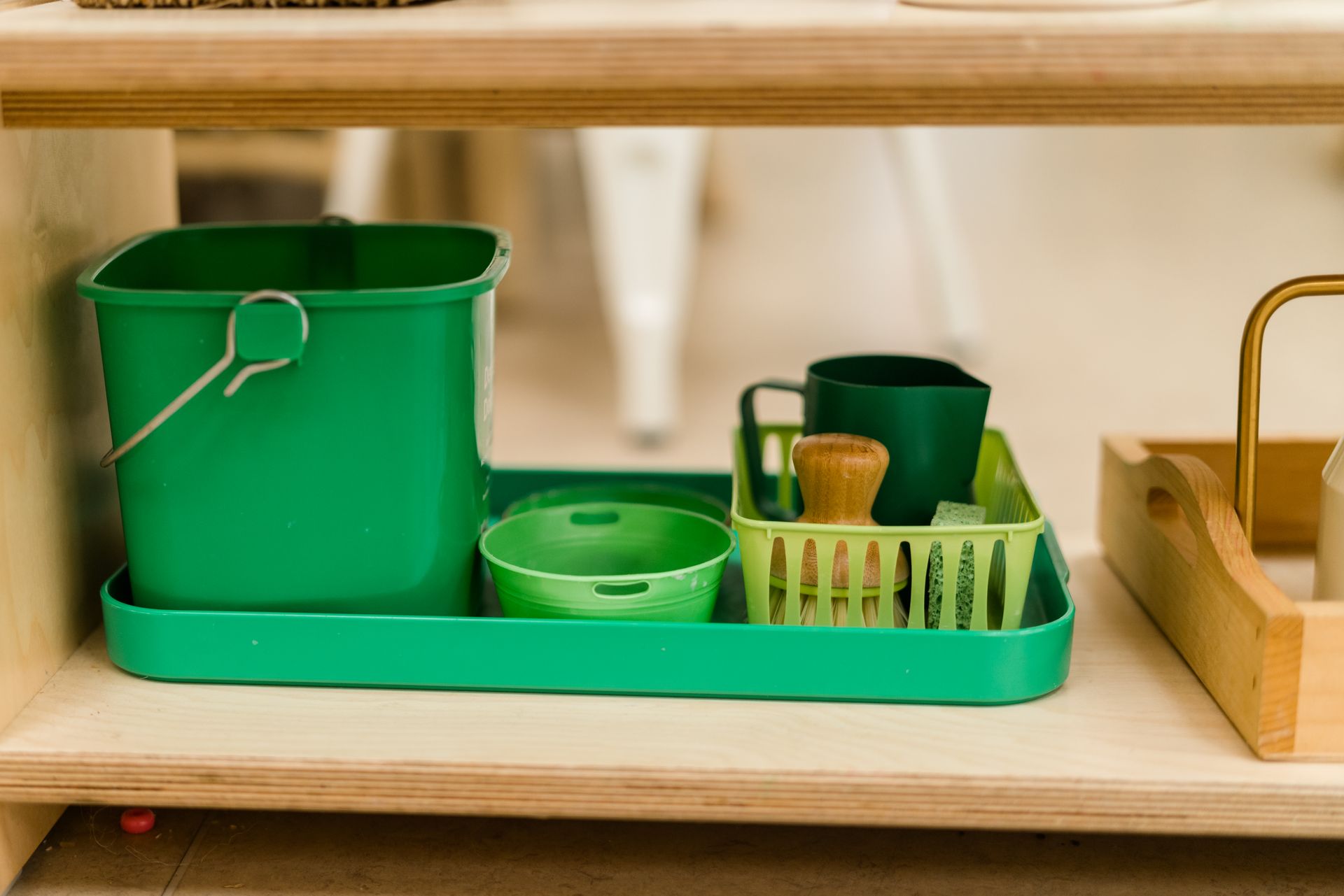 A montessori materials green tray with a bucket, bowls and cups on it is on a wooden shelf.