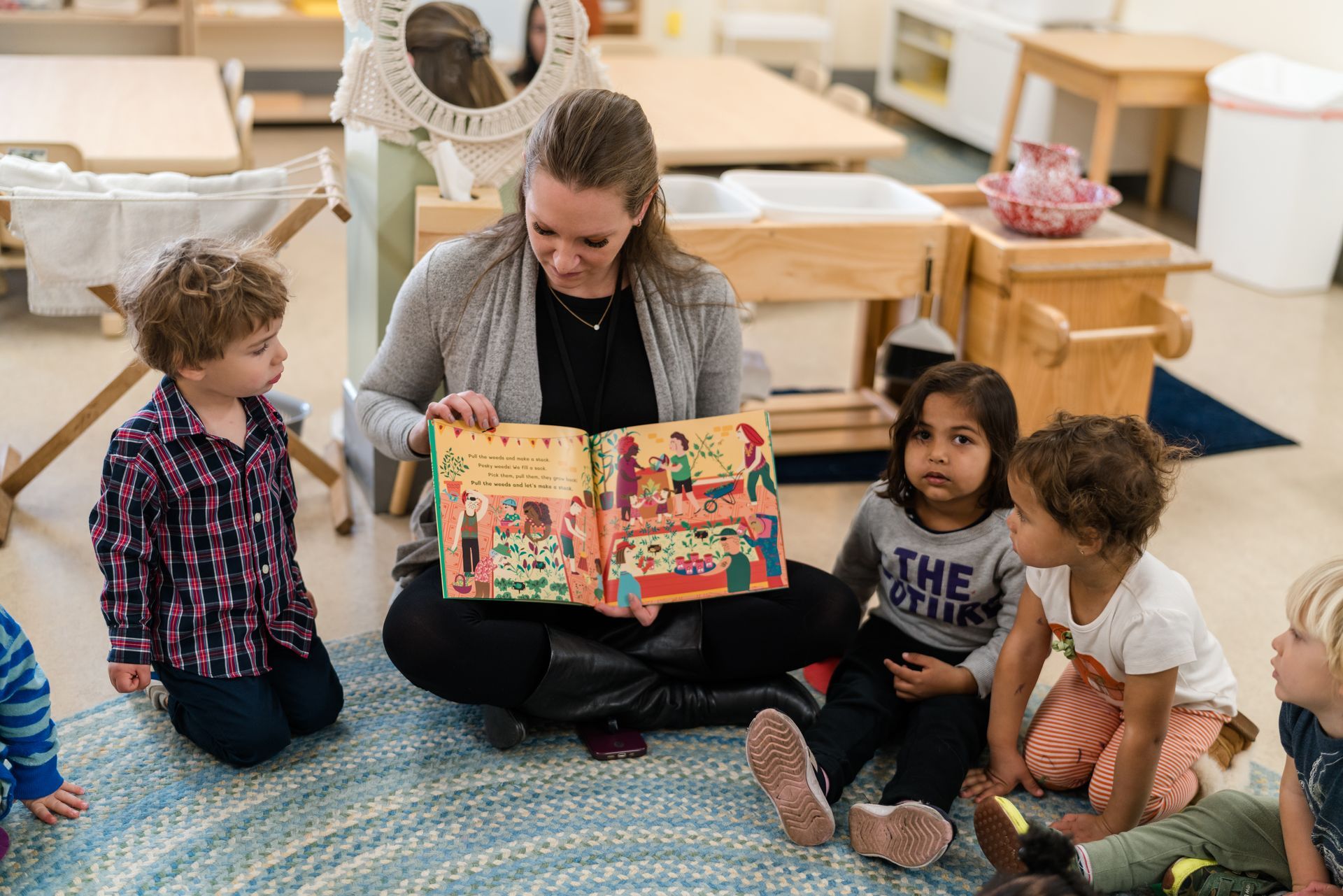 A montessori guide is sitting on the floor reading a book to a group of children.