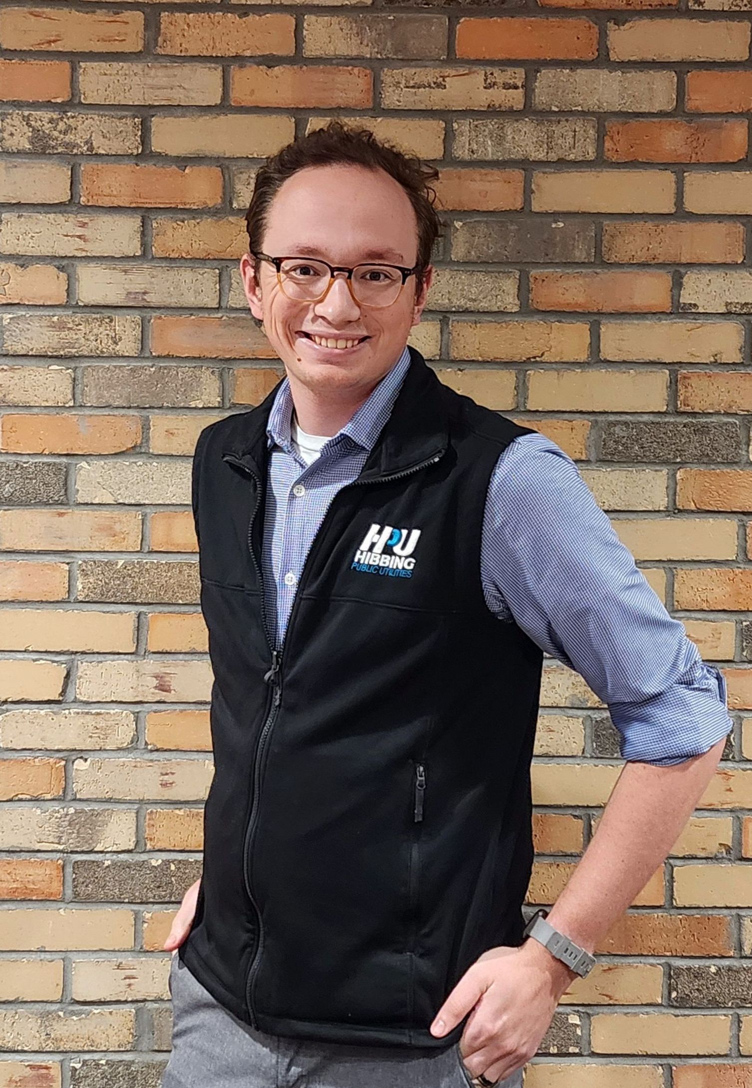 A man wearing glasses and a black vest is standing in front of a brick wall.