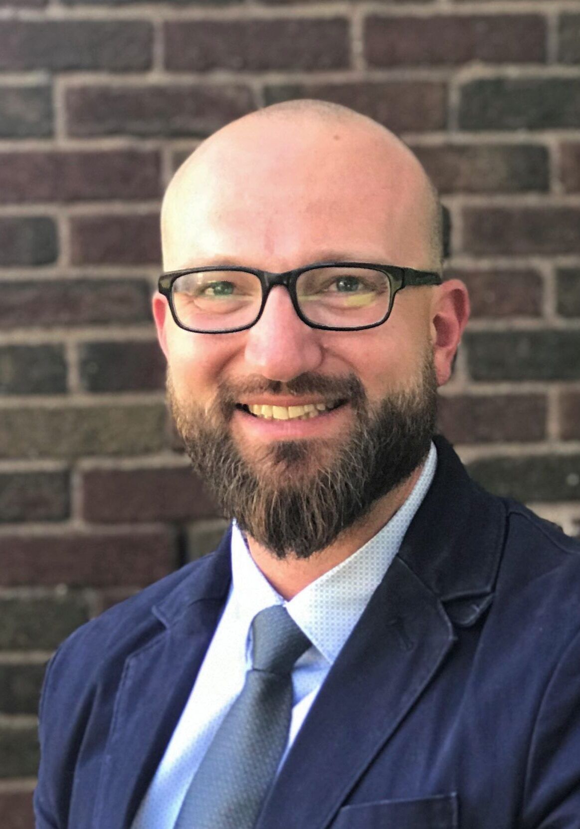A bald man with a beard and glasses is smiling in front of a brick wall.