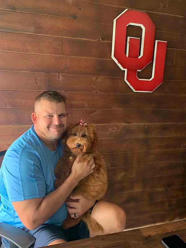 A man is holding a small dog in front of a wooden wall.