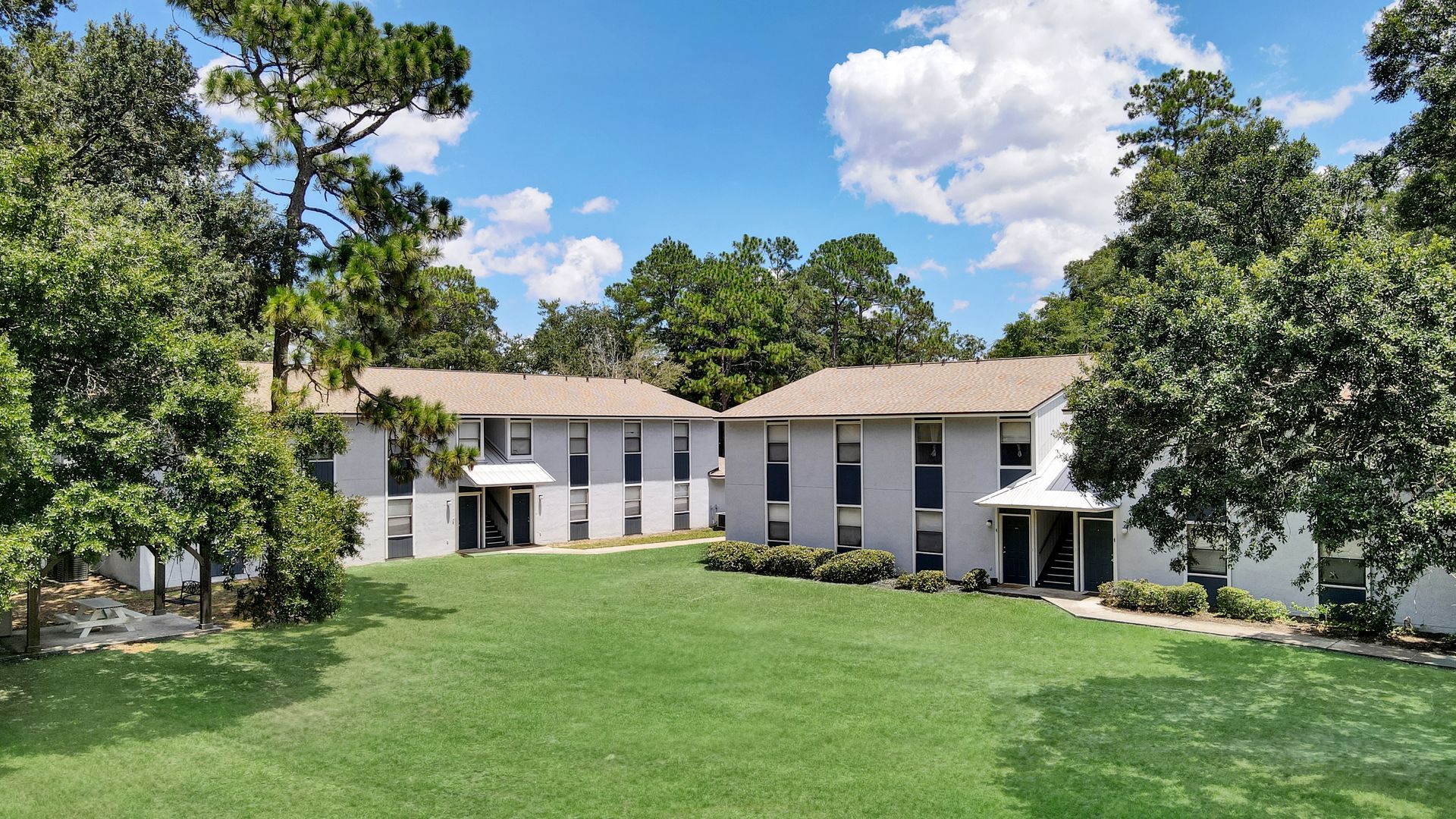Two-story apartment buildings with light gray siding and brown roofs, surrounded by trees and a green lawn under a blue sky.