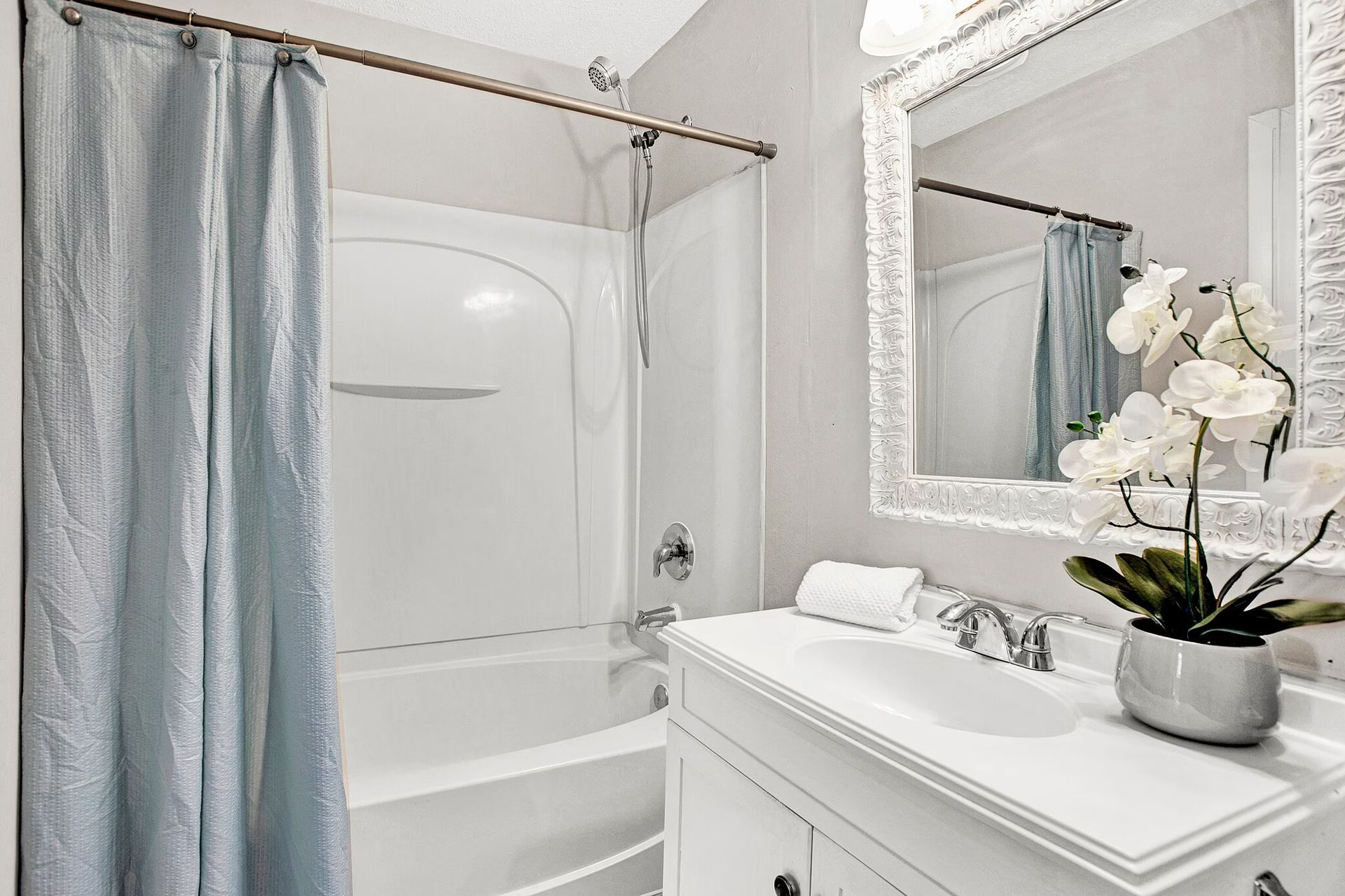 White bathroom with tub, light blue shower curtain, vanity with mirror, and white flowers.