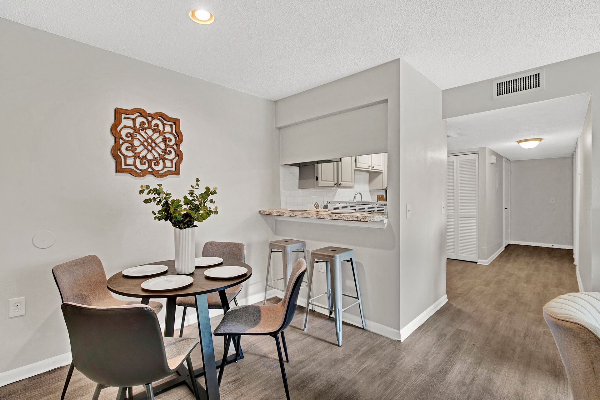 Dining area with a round table, chairs, and a small kitchen bar. Light gray walls and wood-look flooring.