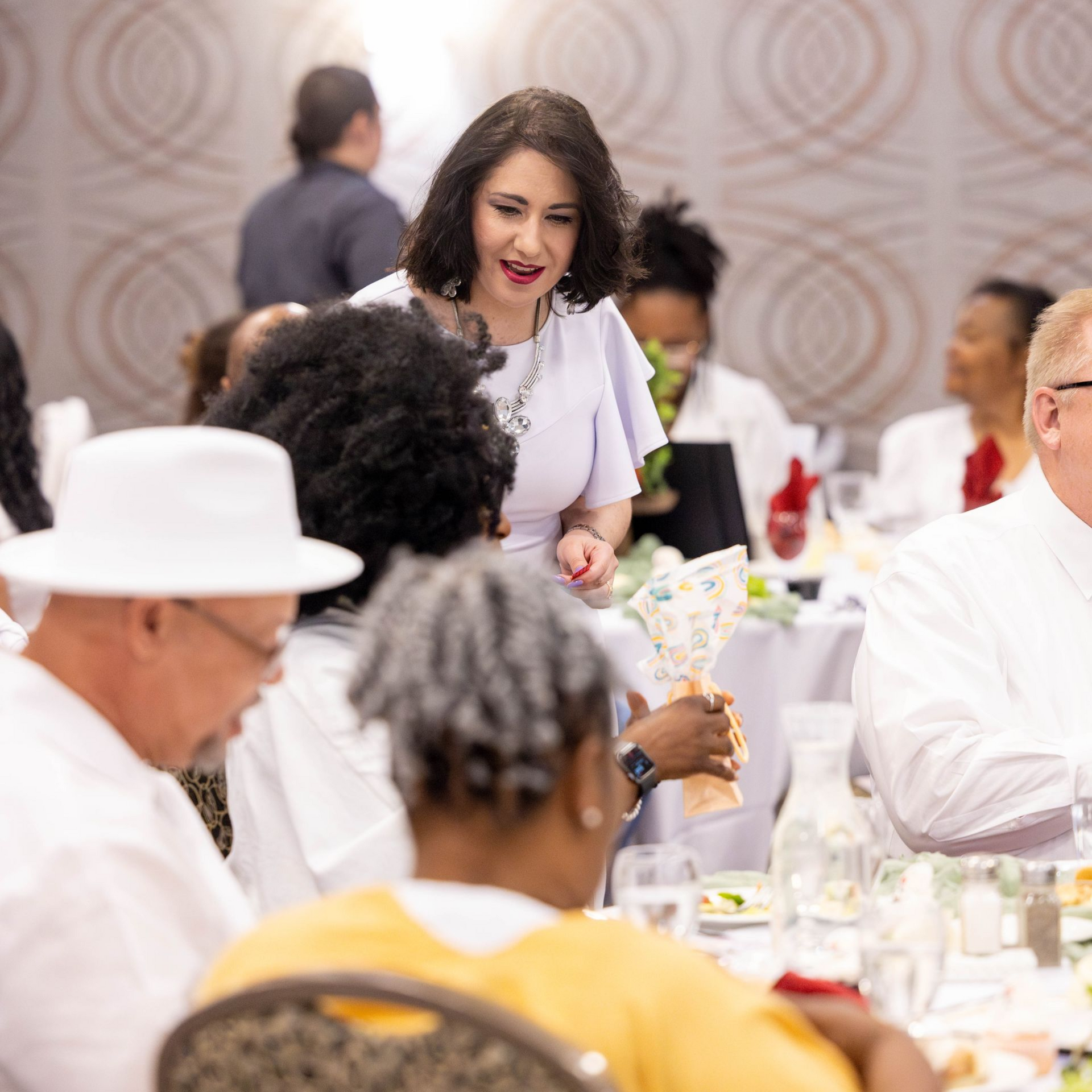 Woman in white dress at a dinner, handing a gift. People seated at tables in a banquet hall.