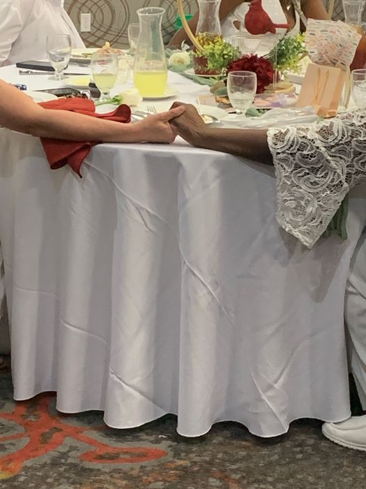 Two people holding hands across a white tablecloth; other diners and food are visible on the table.