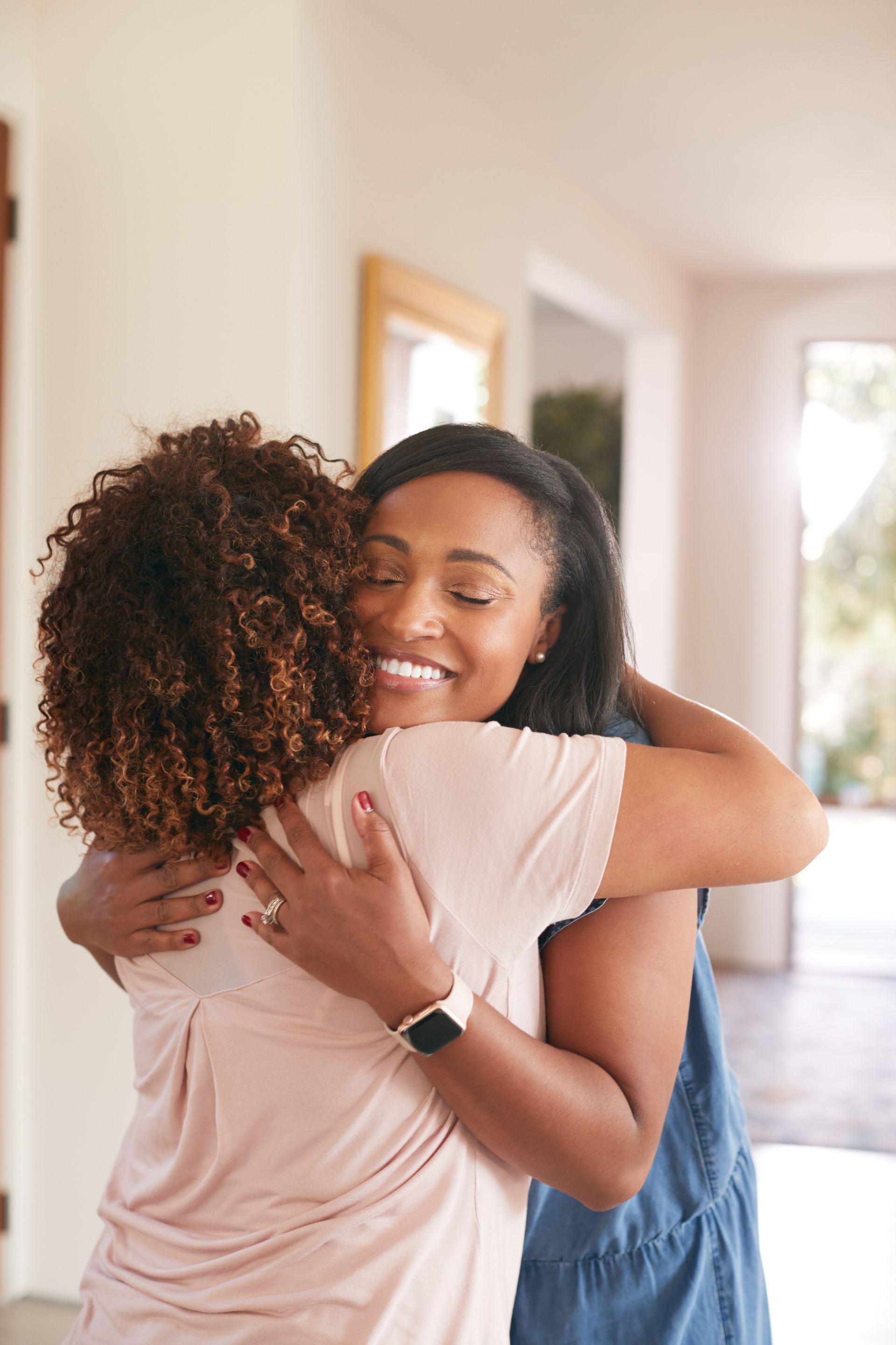 Two women embrace in a well-lit hallway. One smiles warmly, wearing a blue dress and a smartwatch.
