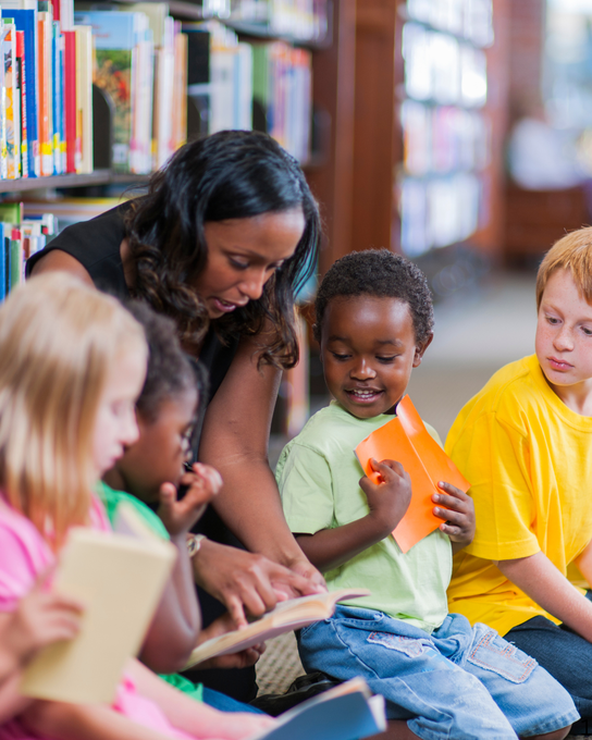 A teacher reads aloud to a diverse group of children holding books in a library.