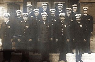 A black-and-white group portrait of sixteen uniformed police officers standing in rows in front of a building.
