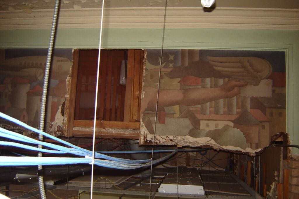 A mural of a hand and buildings behind a broken ceiling in a room with blue hanging cables.
