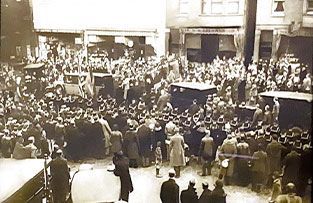 A sepia-toned historical photo of a large, dense crowd of people gathered in a city street near several parked vehicles.