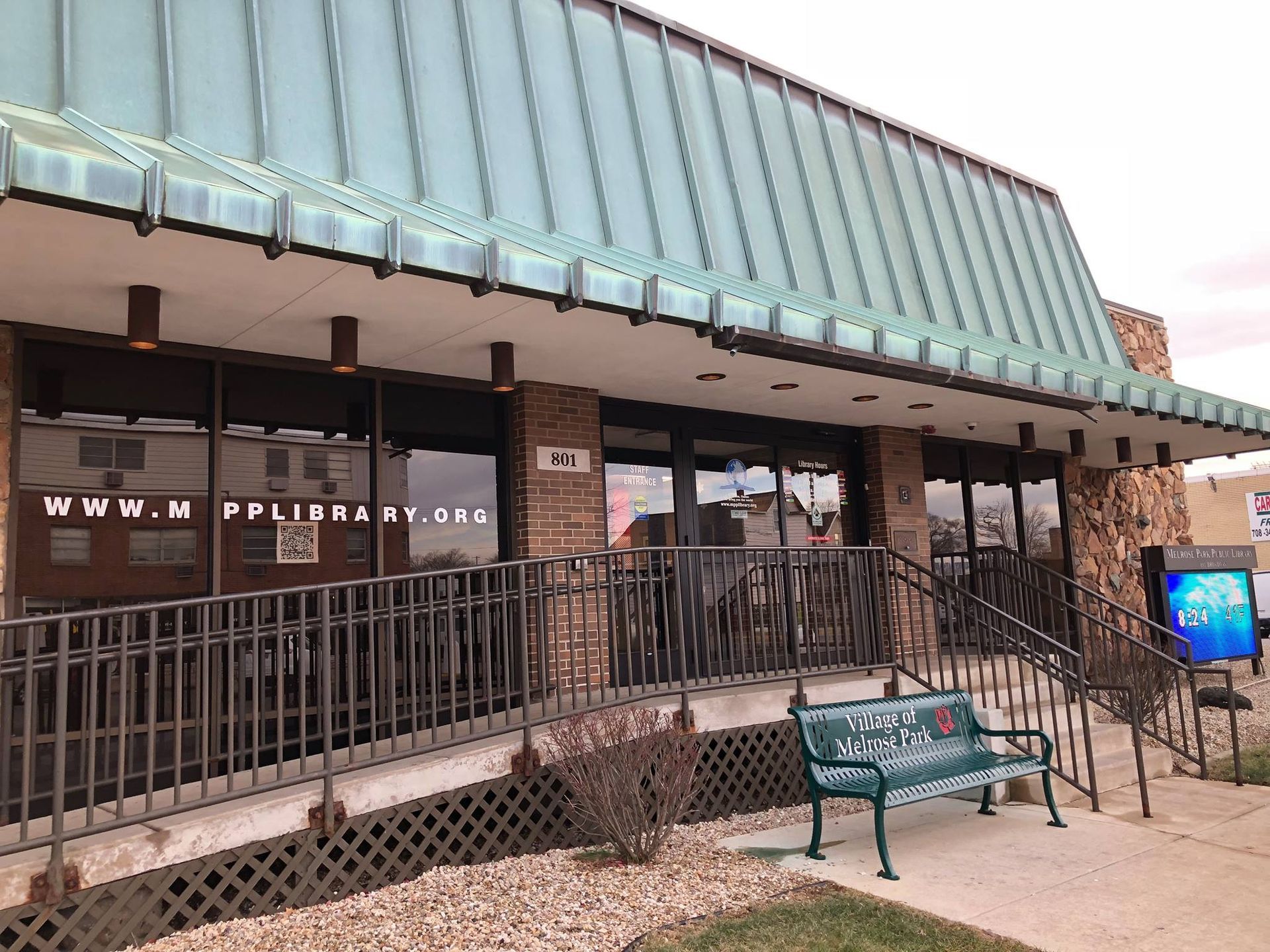 The entrance to the North Platte Public Library, featuring a green awning, brick exterior, metal ramp, and a green bench.