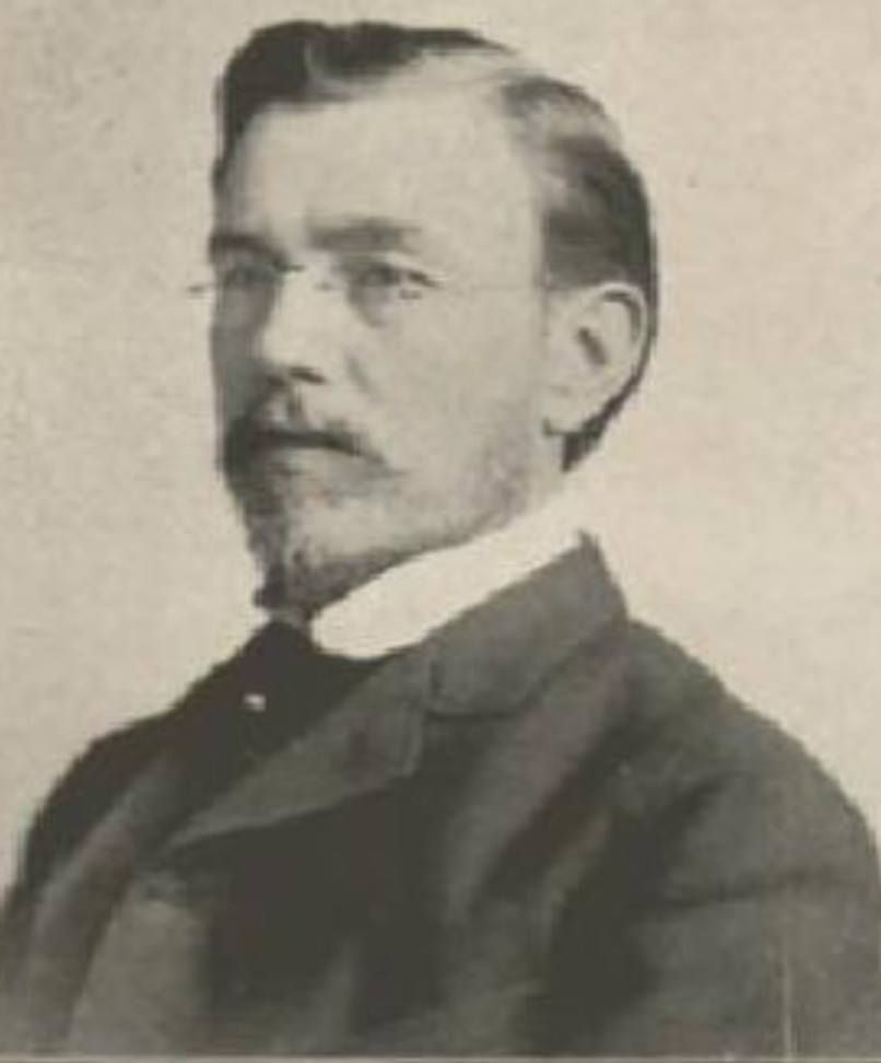 A portrait of a person with a mustache and glasses, wearing a suit jacket and collared shirt.