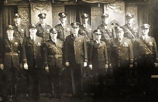 A group of fourteen uniformed police officers stand in two rows for a formal portrait in front of decorative curtains.
