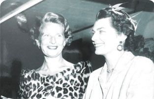 Two women in formal attire smile at each other in a black-and-white portrait.