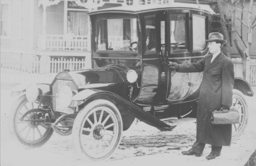 A person in a coat and hat stands next to an early 20th-century enclosed carriage-style automobile, holding a small bag.