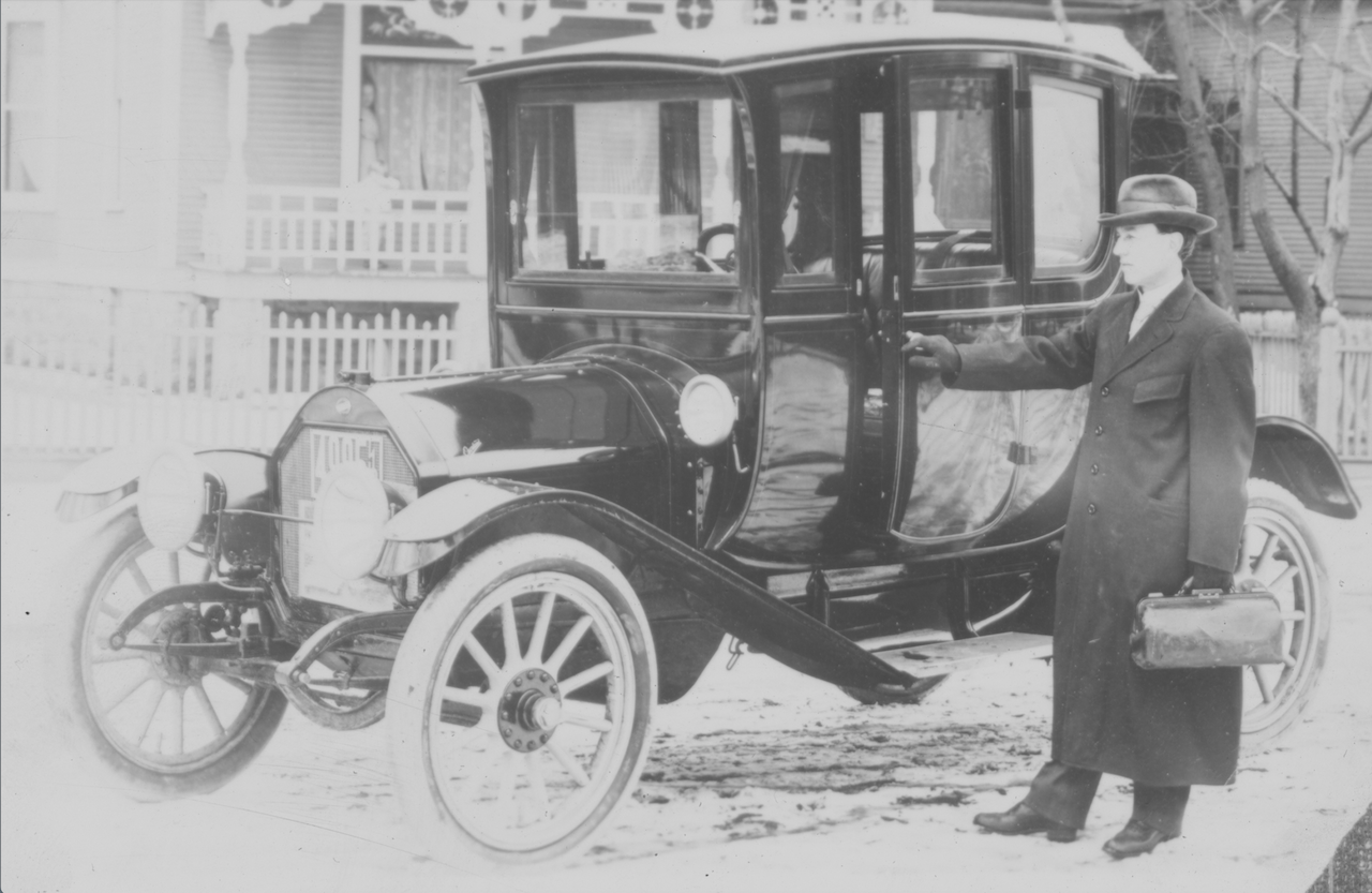 A person in a coat and hat stands next to a vintage black automobile, holding a small bag, in front of a house.