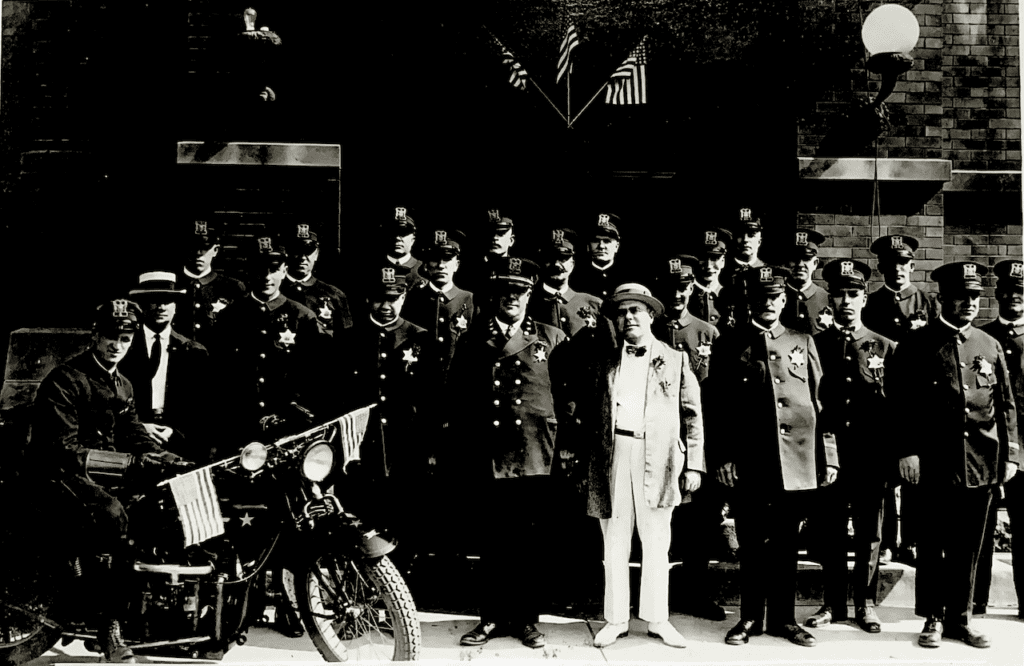 A large group of uniformed police officers stands in front of a building, with one officer on a motorcycle in the front.