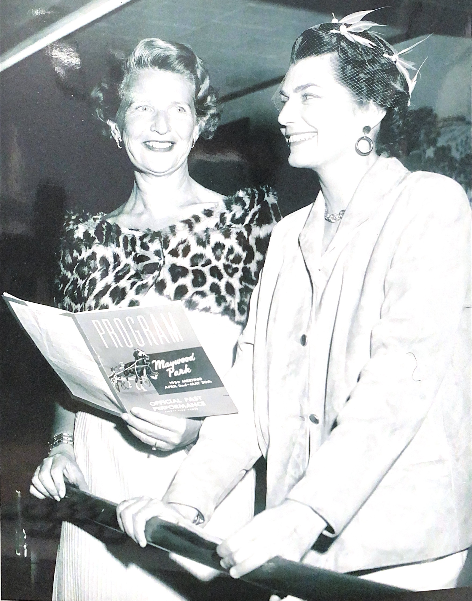 Two people stand at a railing; the person on the left holds a printed program while looking toward the person on the right.