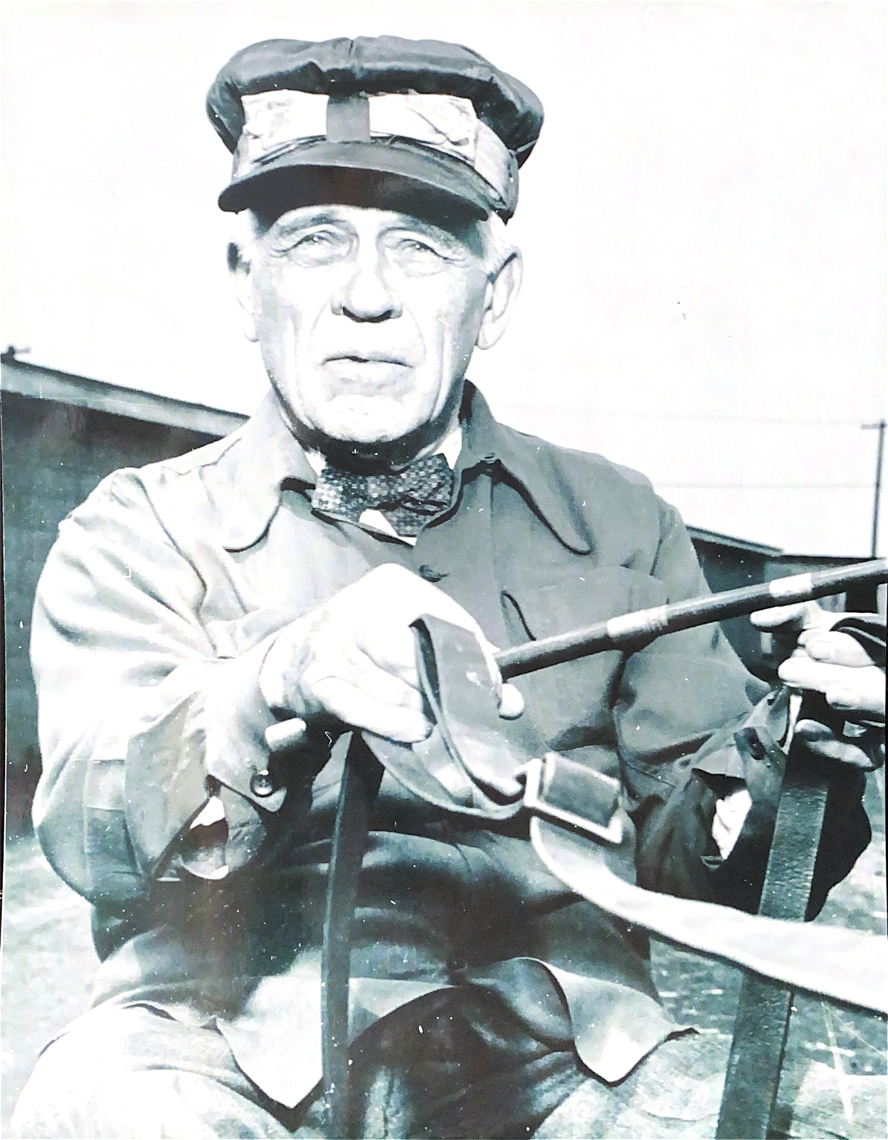 A person wearing a cap, bow tie, and jacket holds a riding crop and leather strap while seated outdoors.