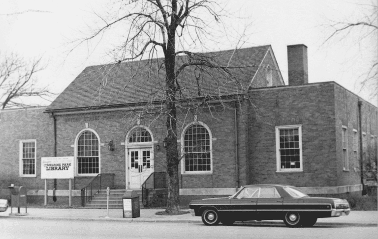 A vintage, black-and-white photo of a brick library building with arched windows and a car parked in front.