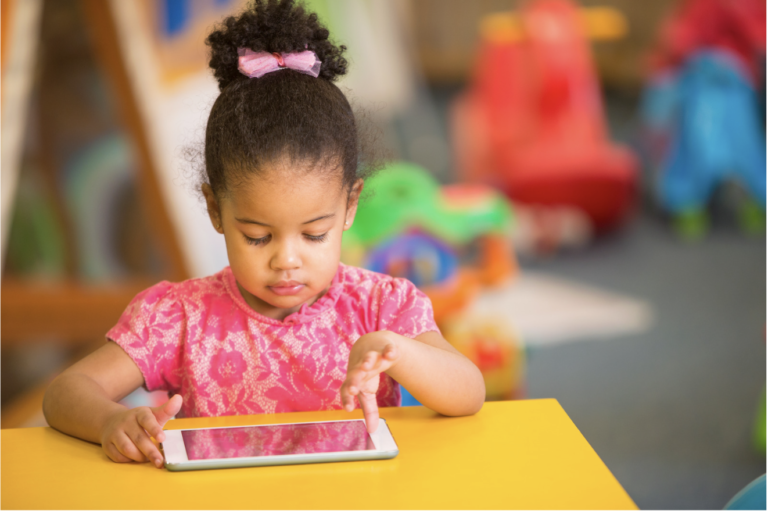 A child in a pink shirt sits at a yellow table, focused on using a tablet screen.